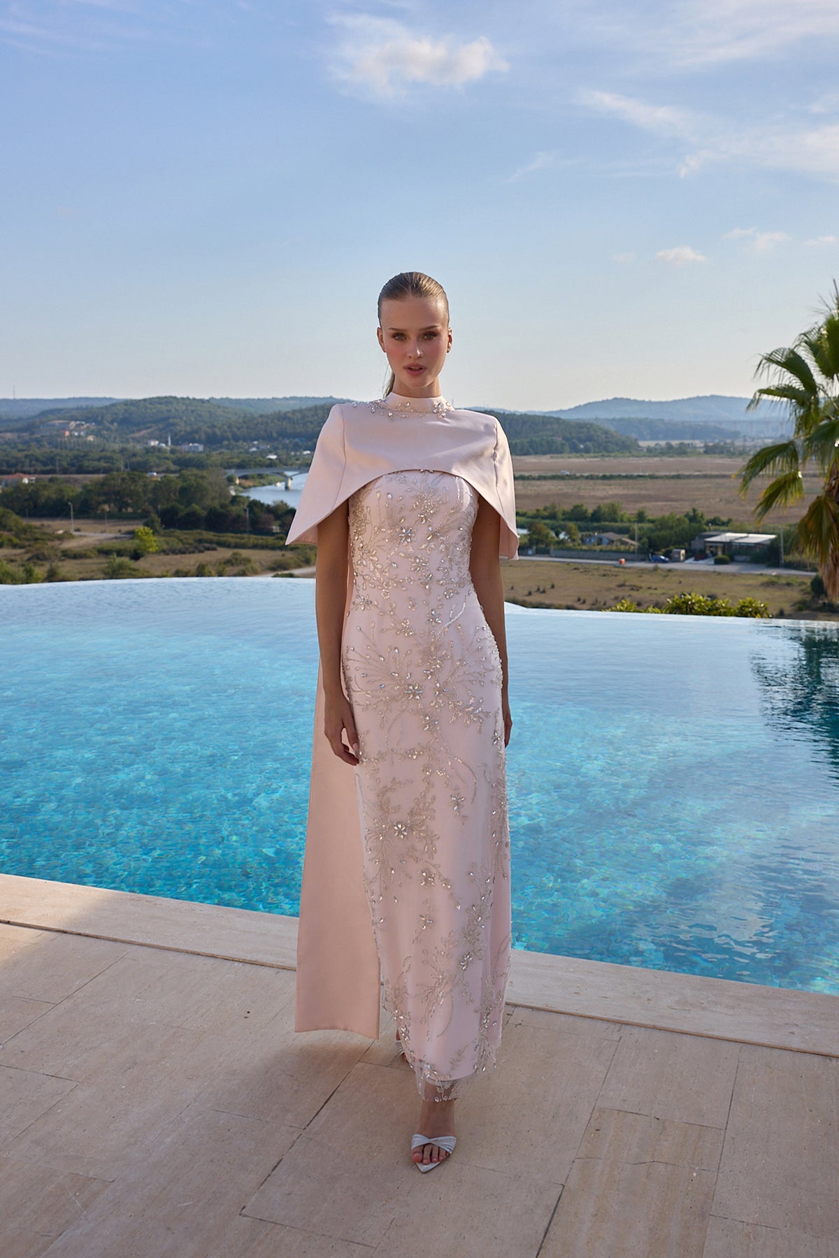 A model stands on a stone terrace with an infinity pool and a scenic landscape in the background, wearing a pale blush pink lace dress with a matching draped cape and silver sandals.