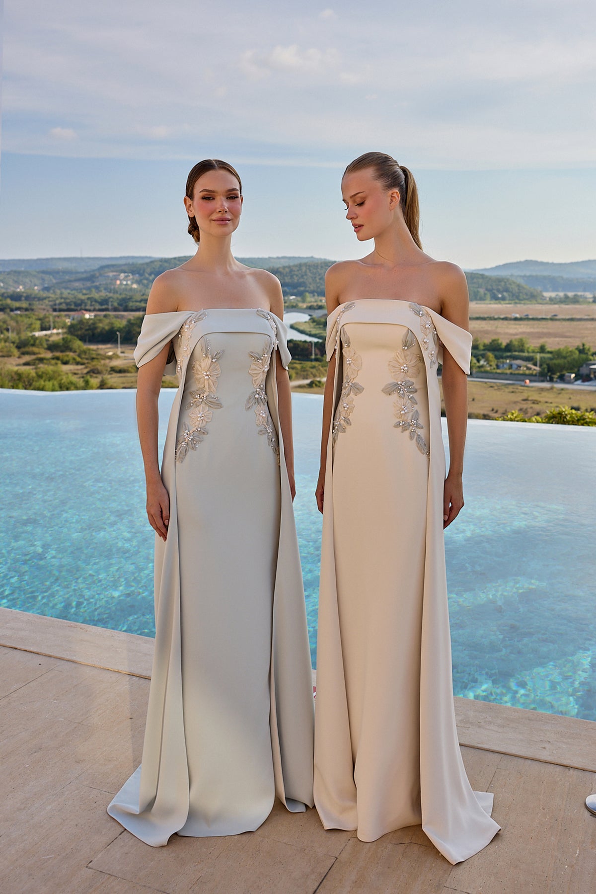 Two women wearing floor-length off-the-shoulder evening gowns with bow details and floral embroidery, standing outdoors by an infinity pool with a scenic landscape in the background.