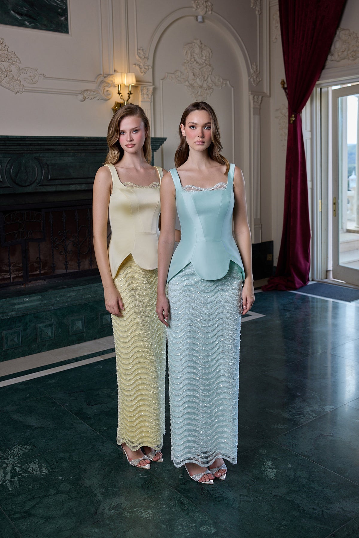 Two women modeling matching sleeveless maxi dresses in an elegant indoor setting with a fireplace and dark marble floor. One dress is pale yellow with a wavy textured skirt, and the other is light blue with a wavy textured skirt.