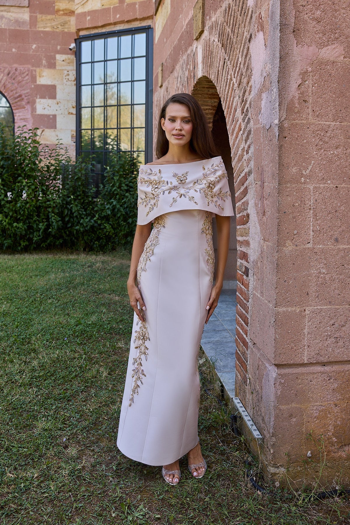 A woman stands outdoors in front of a brick archway wearing a floor-length off-white evening gown with gold embroidery and a draped cap-like yoke. The setting includes grass, brick, and a building facade.