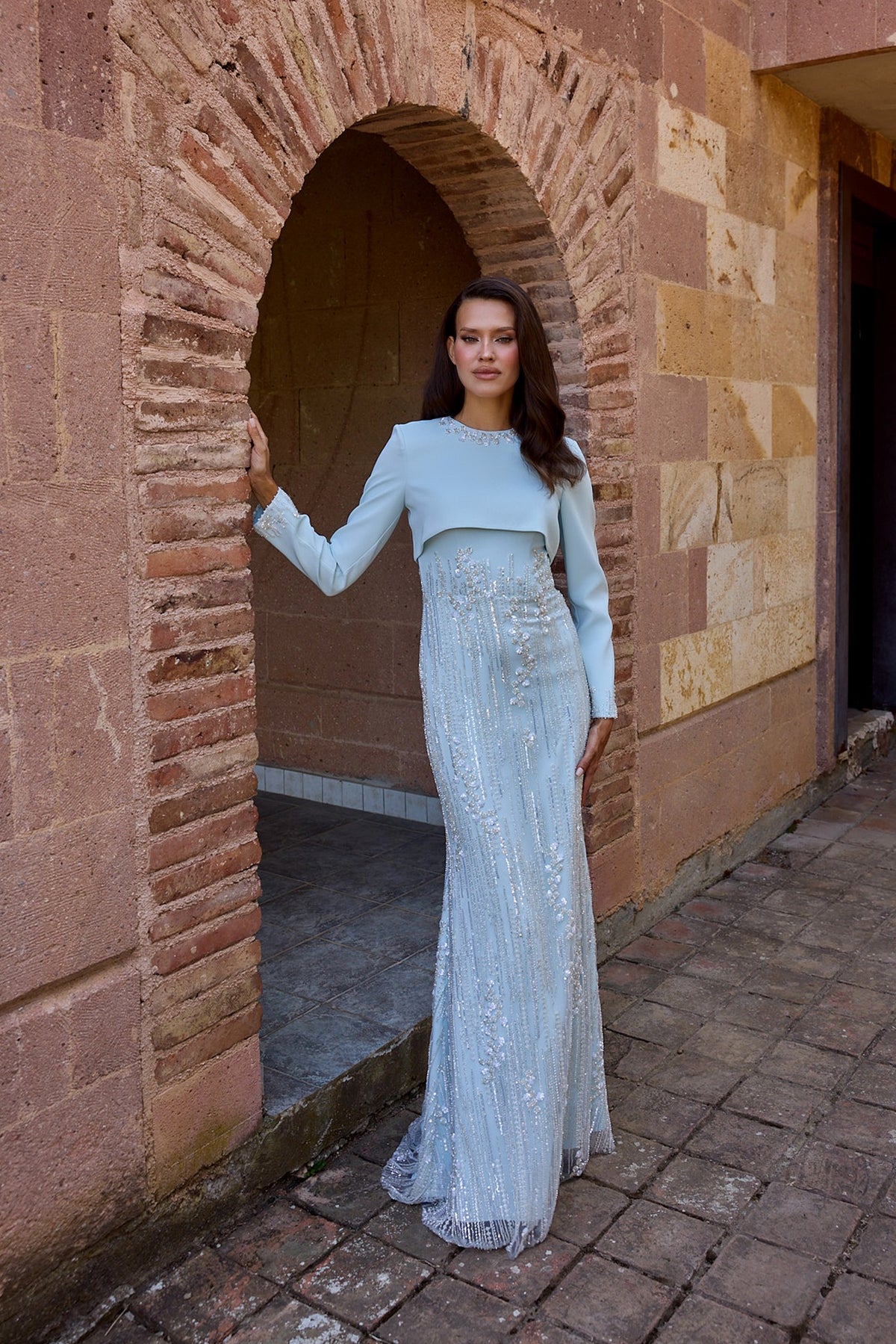 A woman wearing a light blue long-sleeve maxi dress with a cropped top and intricate beadwork, posing in front of a rustic stone archway.