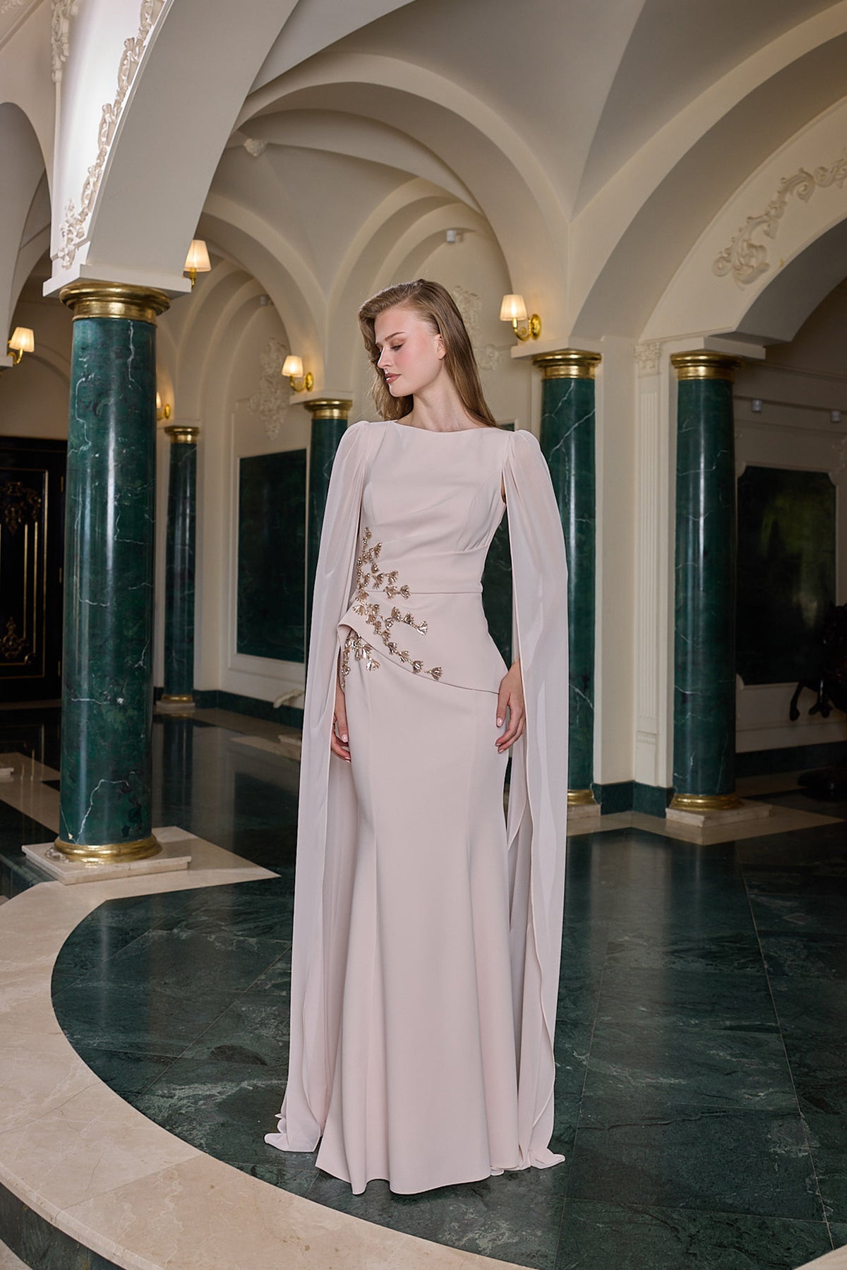 A woman stands in an ornate interior hall wearing a floor-length pale blush gown with draped cape-like sleeves and delicate floral embroidery near the waist. The setting features green marble columns and a dark marble floor.
