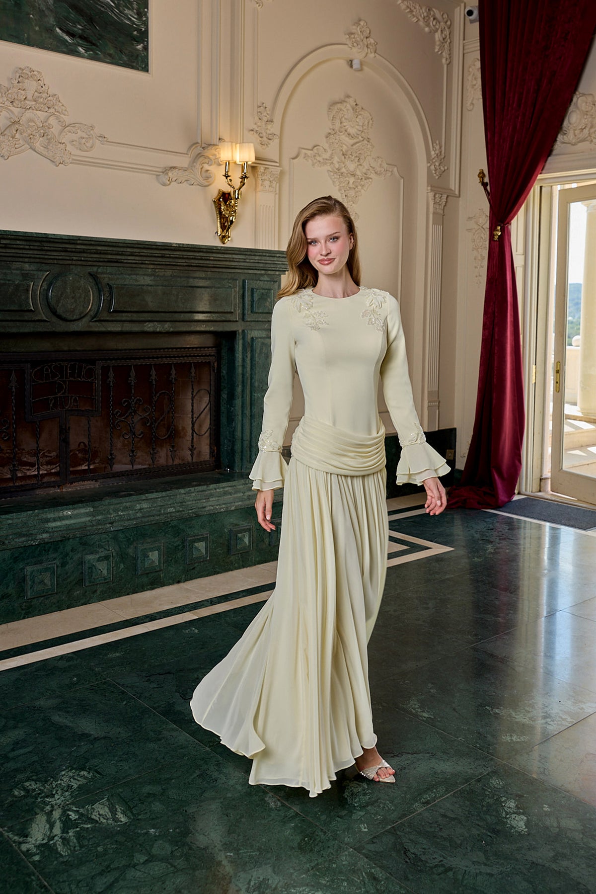 A woman modeling a cream-colored, floor-length evening gown with long flared sleeves and a gathered waist, standing in an elegant room with a dark green marble fireplace and red curtains.