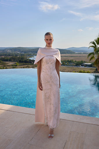 A model stands on a stone terrace with an infinity pool and a scenic landscape in the background, wearing a pale blush pink lace dress with a matching draped cape and silver sandals.