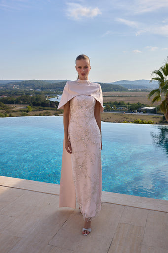 A model stands on a stone terrace with an infinity pool and a scenic landscape in the background, wearing a pale blush pink lace dress with a matching draped cape and silver sandals.