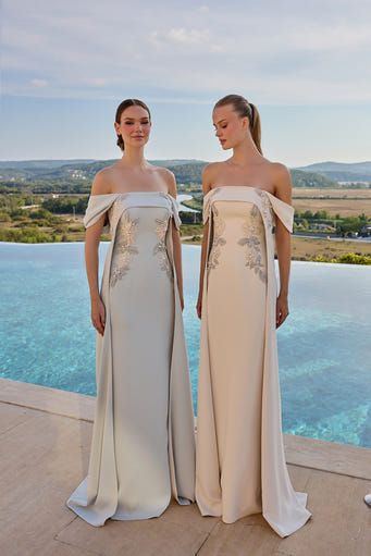 Two women wearing floor-length off-the-shoulder evening gowns with bow details and floral embroidery, standing outdoors by an infinity pool with a scenic landscape in the background.