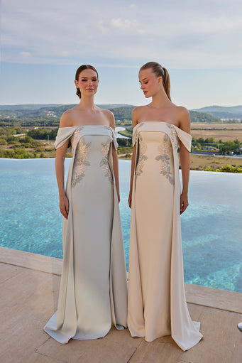 Two women wearing floor-length off-the-shoulder evening gowns with bow details and floral embroidery, standing outdoors by an infinity pool with a scenic landscape in the background.