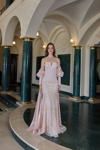 A woman stands in a grand interior hall with arched ceilings and dark green marble columns, wearing a pale blush pink off-the-shoulder evening gown with long puff sleeves and a fitted, embellished mermaid silhouette.