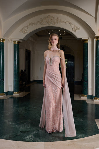 A model stands in an elegant indoor hallway wearing a floor-length pale pink gown with lace texture, a draped neckline, and a sheer draped overlay, set against a classical interior with columns and arches.