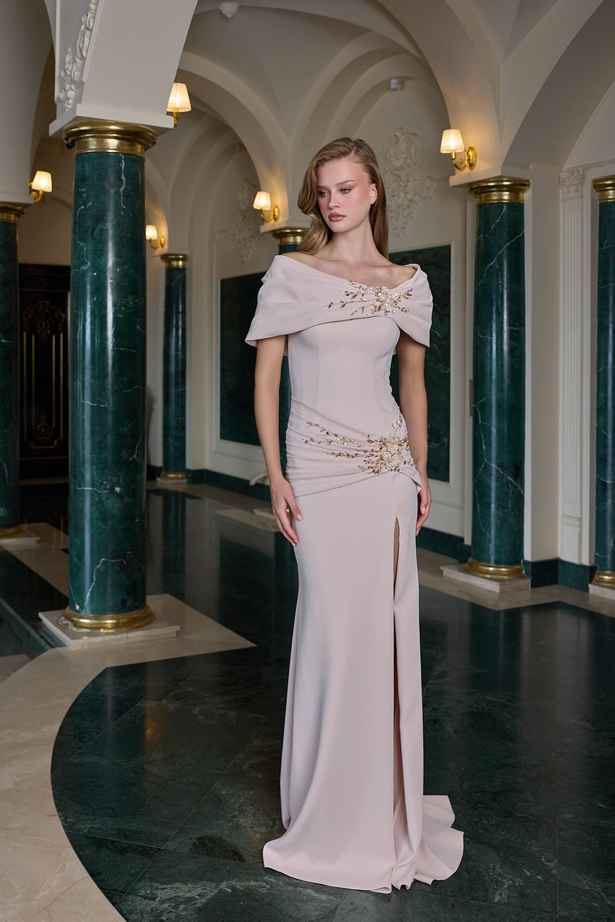 A woman models a floor-length off-the-shoulder evening gown in a grand hall with green marble columns. The gown is a soft blush pink with gold beaded embellishments on the bodice and waist, and a high thigh slit.