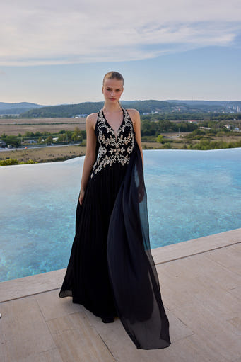 A model stands on a stone terrace beside an infinity pool wearing a sleeveless black evening gown with ornate gold embroidery and a flowing sheer overlay, with a scenic outdoor landscape in the background.