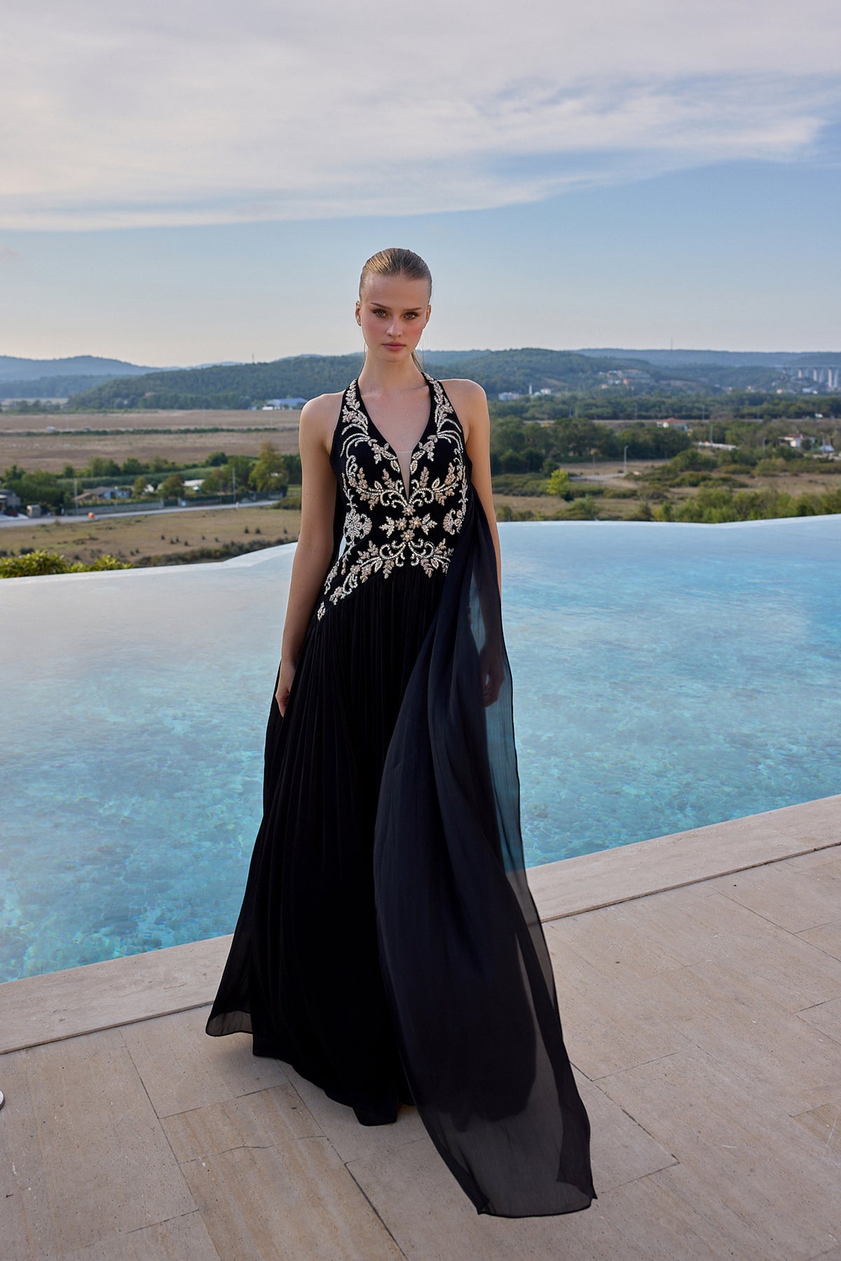 A model stands on a stone terrace beside an infinity pool wearing a sleeveless black evening gown with ornate gold embroidery and a flowing sheer overlay, with a scenic outdoor landscape in the background.