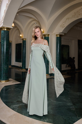 A woman models a floor-length sage green gown with off-the-shoulder lace detailing and a matching lace-trimmed cape, standing in an elegant interior with arched ceilings and dark green marble columns.