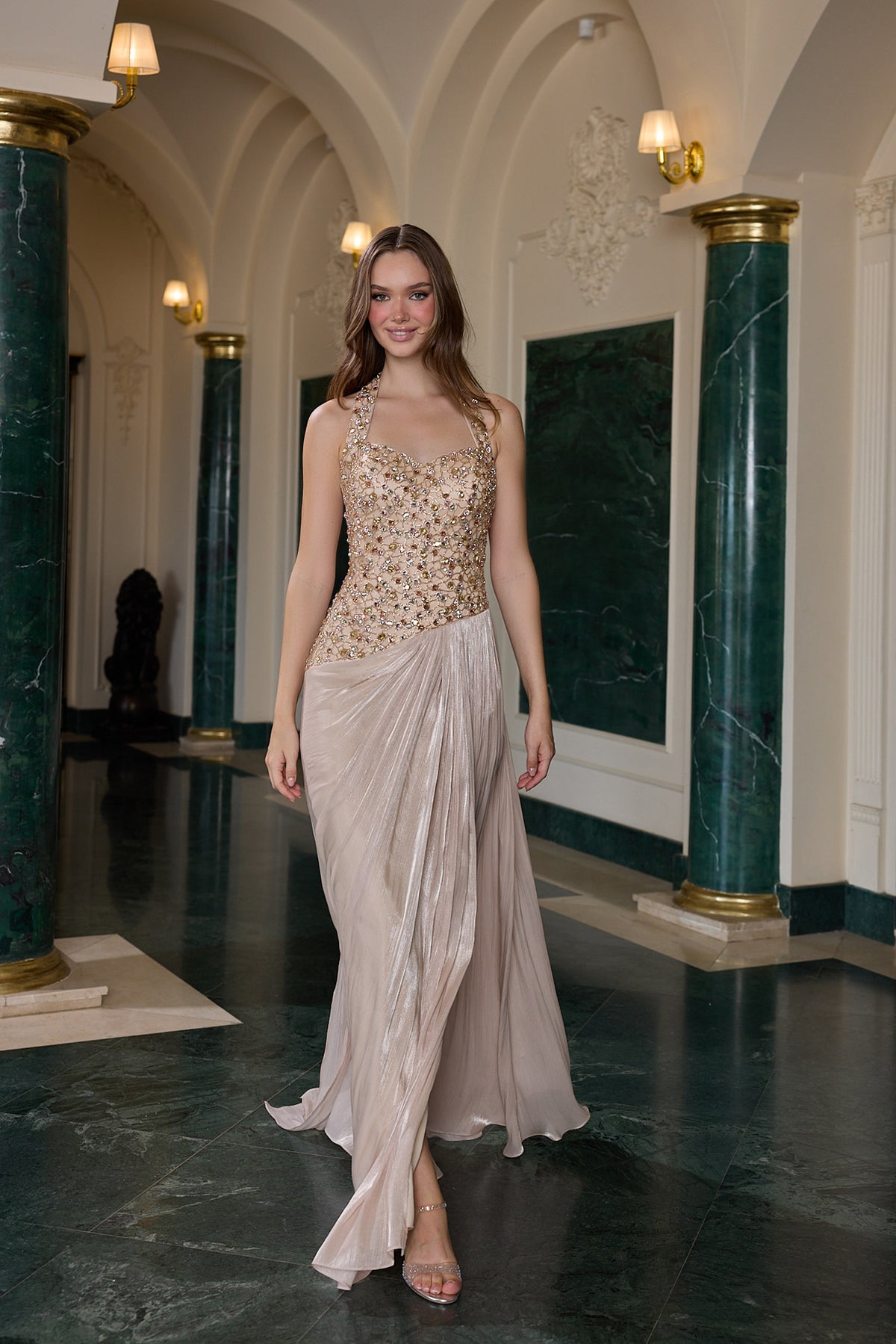A woman models a sleeveless formal gown in a grand hall with green marble columns and arched ceilings. The dress features a fitted bodice covered in gold floral embroidery and a flowing champagne-beige skirt.