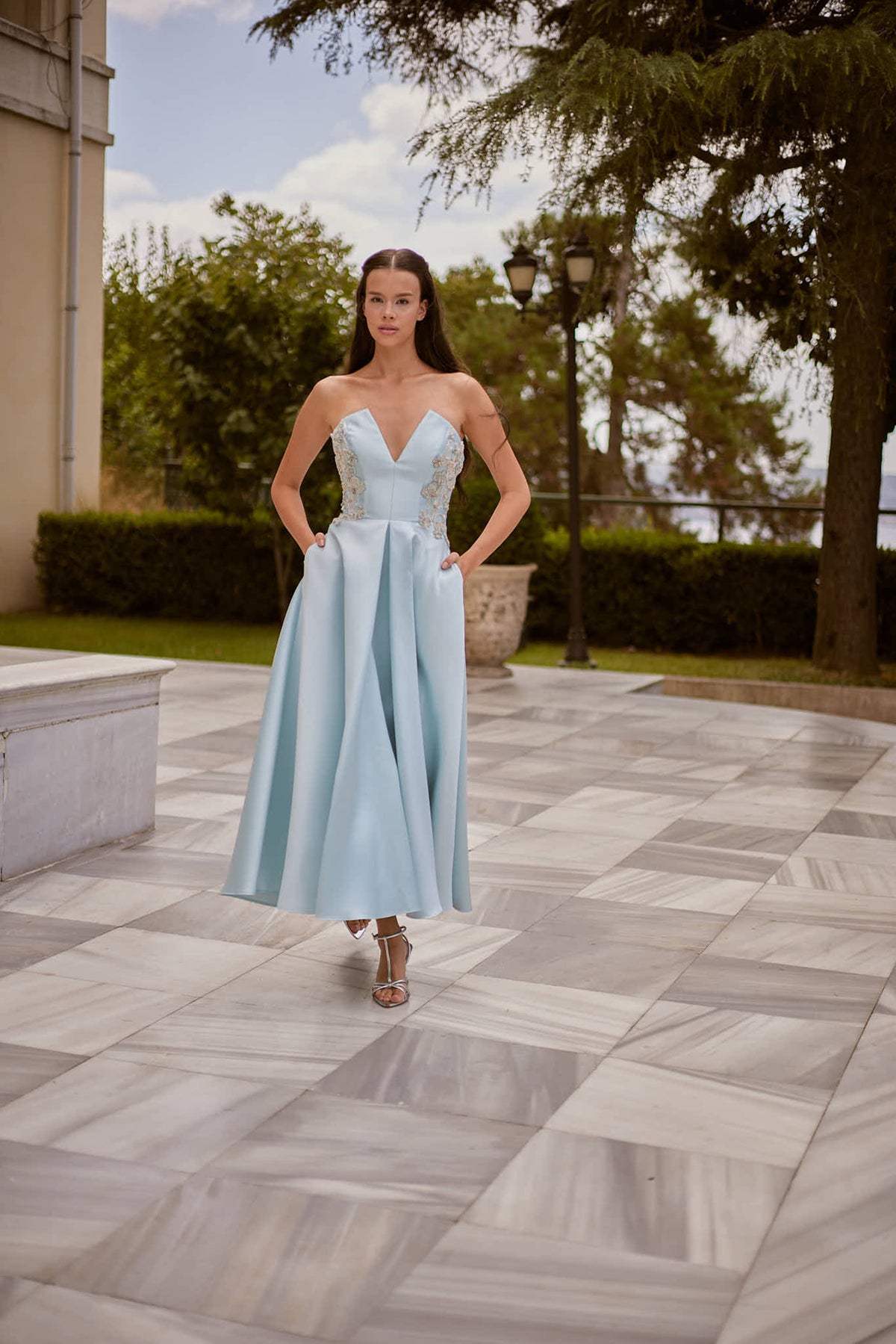 Melisande ice blue satin A-line evening gown, full-length front view with hands in pockets outdoors