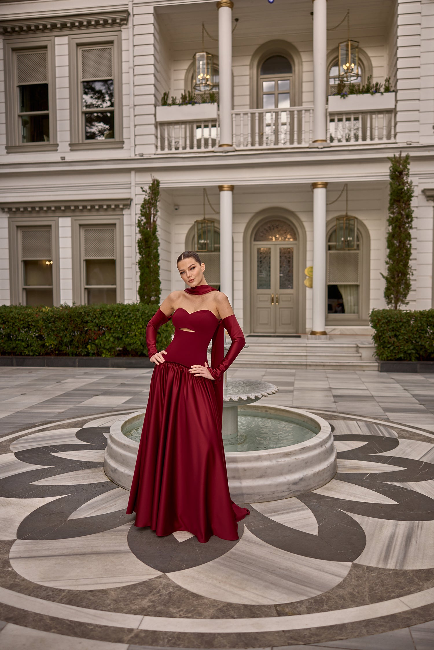 Model wearing Fiorence strapless satin evening gown with matching gloves and scarf detail in front of a luxury mansion