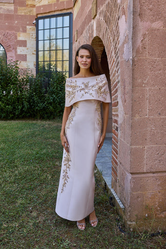 A woman stands outdoors in front of a brick archway wearing a floor-length off-white evening gown with gold embroidery and a draped cap-like yoke. The setting includes grass, brick, and a building facade.