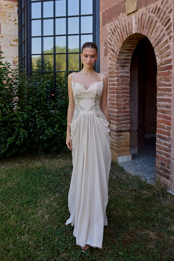 A woman wearing a floor-length ivory wedding gown stands outdoors on grass beside a brick archway and large black-framed window. The dress has thin straps, a fitted embellished bodice, and a flowing skirt.