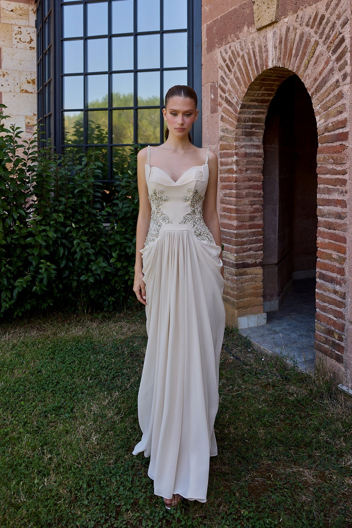 A woman wearing a floor-length ivory wedding gown stands outdoors on grass beside a brick archway and large black-framed window. The dress has thin straps, a fitted embellished bodice, and a flowing skirt.
