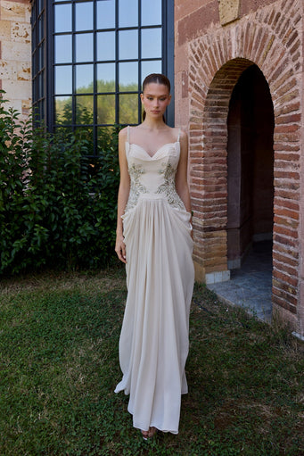 A woman wearing a floor-length ivory wedding gown stands outdoors on grass beside a brick archway and large black-framed window. The dress has thin straps, a fitted embellished bodice, and a flowing skirt.