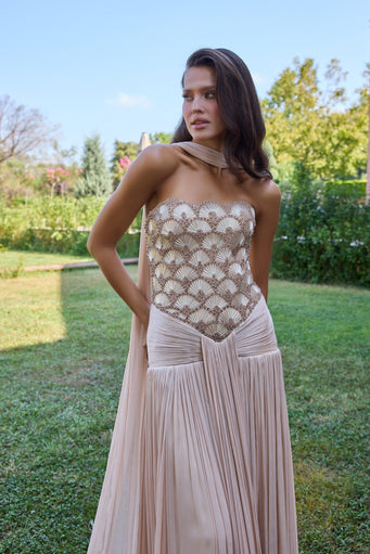 A woman wearing a strapless champagne lace corset top with a matching pleated blush pink skirt, standing outdoors on a grassy lawn with trees and blue sky in the background.
