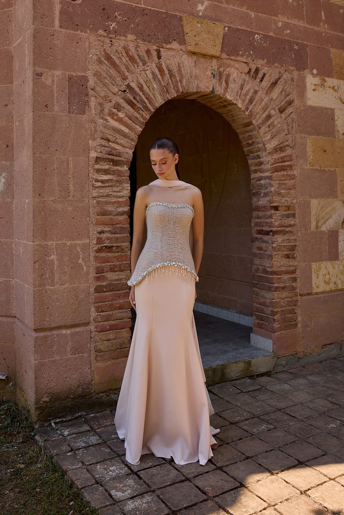 A woman wearing a champagne strapless gown with a beaded corset-style bodice and a flowing satin mermaid skirt, standing in front of a rustic stone archway.