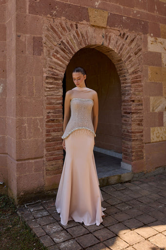 A woman wearing a champagne strapless gown with a beaded corset-style bodice and a flowing satin mermaid skirt, standing in front of a rustic stone archway.