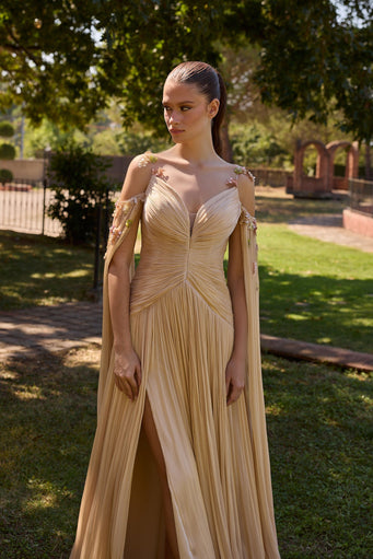 A woman wearing a floor-length champagne beige gown with draped cape-like sleeves and a plunging neckline, standing outdoors in a garden setting with trees and grass in the background.